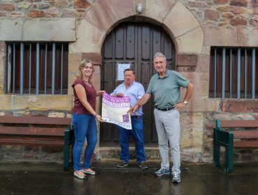 El concejal de Festejos, Fernando Sañudo (en el centro), sostiene el cartel de la fiestas de Posadillo junto a la ermita del pueblo, acompañado de los concejales Avelino Rodríguez y María José Alvarez